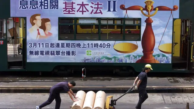Workers move supplies past a tram decorated with an advertisement encouraging Hong Kong people to increase their knowledge of the Basic Law, the territory's constitution, 07 May 1999 in Hong Kong. The government has been fiercely criticized by lawmakers for compremising the Basic Law and the independence of the judiciary by being non-committal when asked whether it would implement a high court ruling which gives the right of abode to some 1.67 million mainland born children of Hong Kong parents. The government has suggested that it may ask Beijing's national parliament to re-interpret the law, a move that would jeopardize Hong Kong's autonomy. (ELECTRONIC IMAGE) AFP PHOTO/Robyn BECK (Photo by ROBYN BECK / AFP) (Photo by ROBYN BECK/AFP via Getty Images)