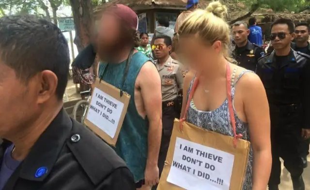 Homem e mulher escoltados desfilando com cartaz no pescoço