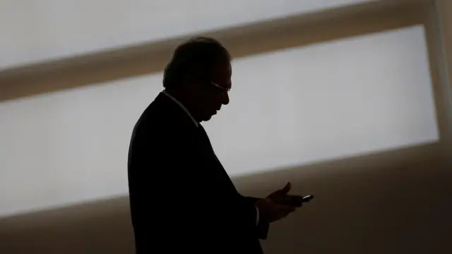 Paulo Guedes olha para celular em ambiente interno