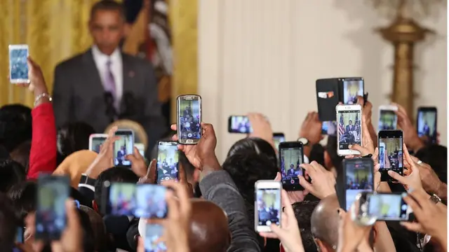 Decenas de celulares toman fotografías de Obama quien aparece en el fondo de manera borrosa.