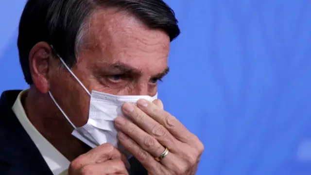 Em frente a painel azul, Bolsonaro apareceslowroll pokerlado ajeitando máscara