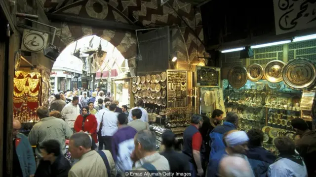 Dezenas de pessoas andam em corredor de mercado, de onde se vê colares e artefatos de cozinha à venda