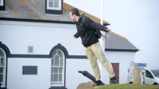 Мужчина пытается устоять на ногах под порывами ветра в Лендс-Энде