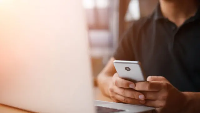 Joven frente a su computadora con un celular en la mano