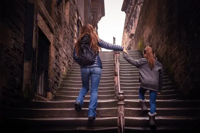 mother and her daughter. They are holding hands and walking up an old stone staircase between the gothic buildings of Edinburgh.
