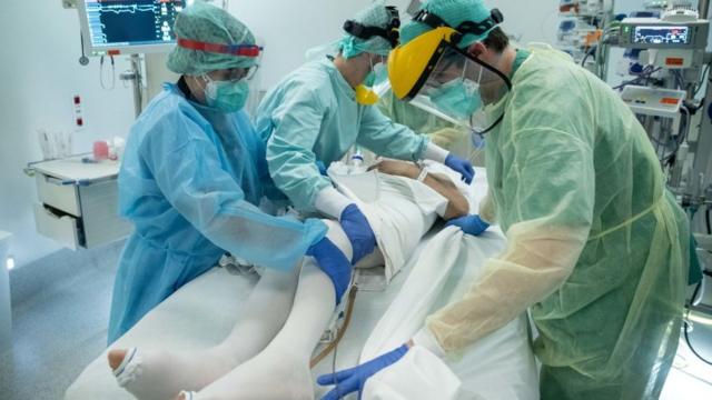 Três profissionais de saúde em uma sala de terapia intensiva, atendendo um paciente de covid-19