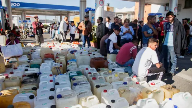 Galones acumulados en el piso, al lado de personas que esperan conseguir gasolina.