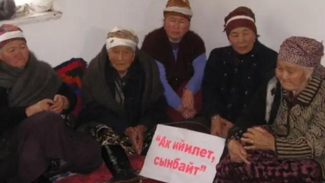 Исаковдун туугандарынан үч-төрт улгайган аялдар дагы моюнуна "Ак ийилет, бирок сынбайт" деген жазууну илип алып, жуурканга оронуп олтурушуптур.