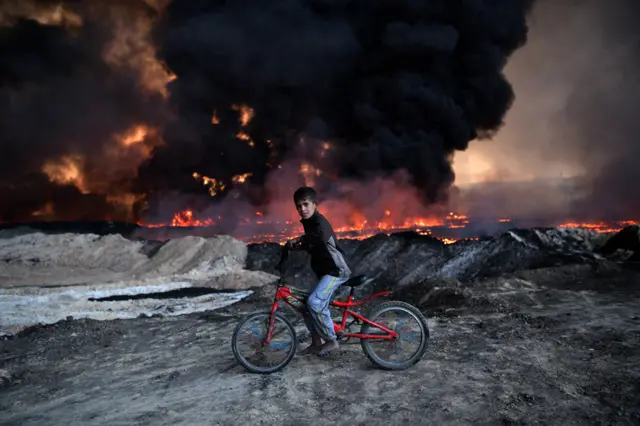 A boy pauses on his bicycle as he passes an oil field that was set on fire by retreating ISIS fighters ahead of the Mosul offensive, on 21 October 2016 in Qayyarah, Iraq.