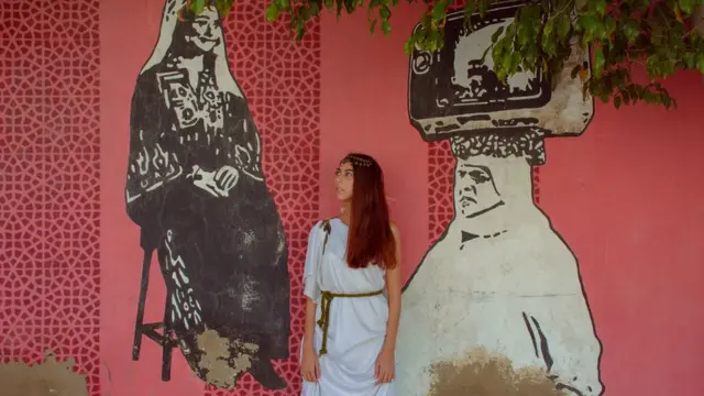 شابة ترتدي زي روماني بجانب رسم لامرأة تحمل تليفزيون على رأسها ورسم آخر لفلاحة