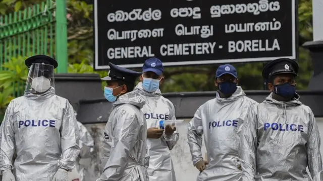 Police personnel stand guard as protesters hold placards during a demonstration against the government policy of forced cremations of Muslims who died of the Covid-19 coronavirus, outside a cemetery in Colombo on December 31, 2020. (Photo by ISHARA S. KODIKARA / AFP) (Photo by ISHARA S. KODIKARA/AFP via Getty Images)