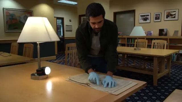 Franco Galvão em pé, debruçado diante de mesa, manipulando partituras com luvas em ambiente de biblioteca