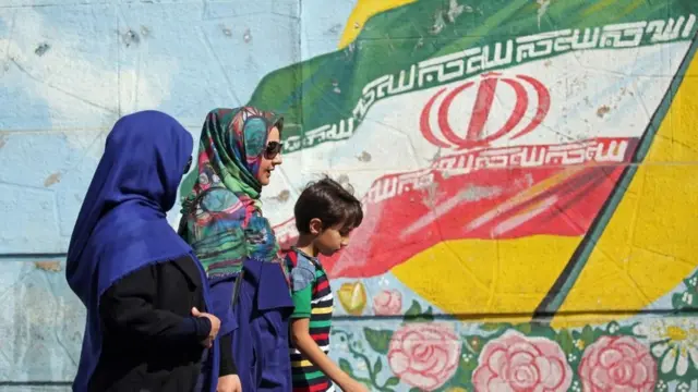 Dos mujeres y un niño caminan delante de un mural con la bandera iraní.