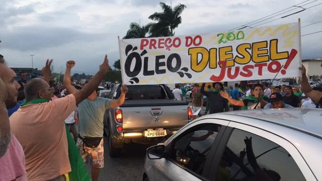 Manifestantes na porta de refinaria, ao lado de cartaz que pede: 'Preço do óleo diesel justo!'