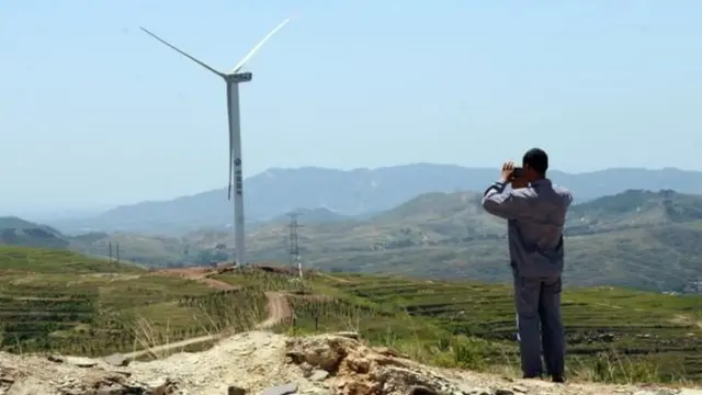 Un habitante de un poblado rural en China mira el horizonte con unos binoculares. Al fondo, un molino que produce electricidad por viento.