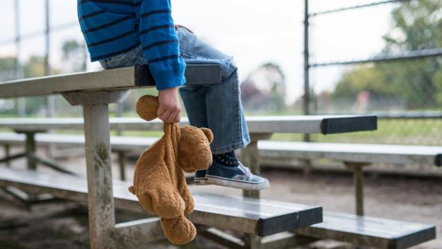 Foto ilustrativa sobre abuso infantil - menino sentado em arquibancada com ursinho de pelúcia