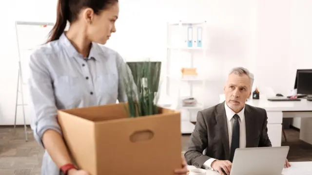 Una mujer con una caja con pertenencias mira de reojo a un hombre sentado en un escritorio.