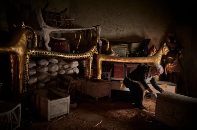 Темна кімната з дерев'яними предметами і скриньками і чоловік в окулярах