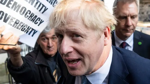 Boris Johnson disambut pemrotes yang membawa poster bertuliskan antara lain Demokrasi dan Kedaulatan di depan kantornya di London, Selasa (23/07).