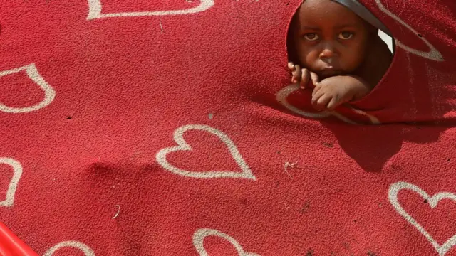 A child looks at the camera through a hole in a blanket which is attached to a temporary shelter.