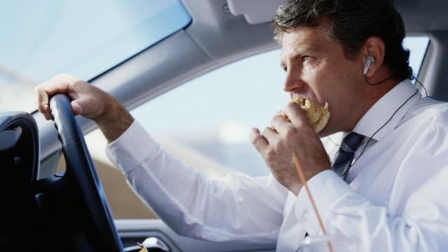 Un hombre conduce un auto mientras come una hamburguesa y escucha por un audífono