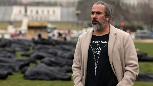 Detrás de Manuel Oliver se ve la instalación de 1.100 bolsas para cadáveres que deletrean las palabras "thoughts and prayers" (pensamientos y oraciones), una frase común de los políticos estadounidenses tras los tiroteos.