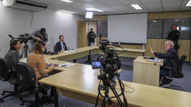 A equipe da BBC e o ex-presidente Lula em mesas, observados por agente de segurança
