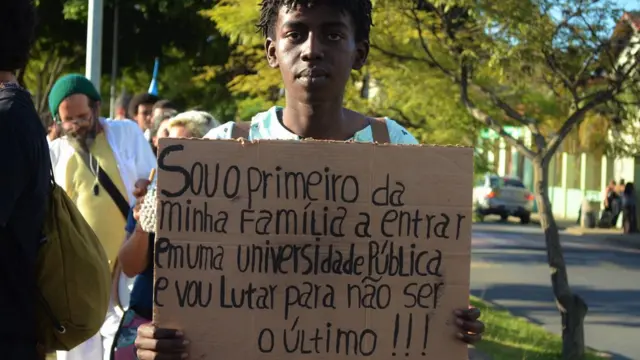 Jovem negro segurando um cartaz que diz 'Sou o primeiro da minha família a entrarapostar cassinouma universidade pública e vou lutar para não ser o último'