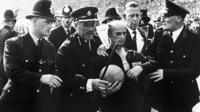 A referee being escorted from the pitch by policemen