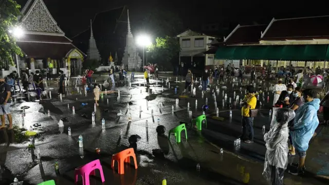 ประชาชนจำนวนมากไปต่อแถวเพื่อรอรับบัตรคิวเข้าตรวจเชิงรุกหาเชื้อโควิด-19 ท่ามกลางสายฝนที่ตกลงมาอย่างหนัก โดยแต่ละคนได้วางขวดน้ำและของใช้แทนตัวเองระหว่างหลบฝน ที่วัดบางขุนเทียนนอก เขตจอมทอง เมื่อ 9 ก.ค.