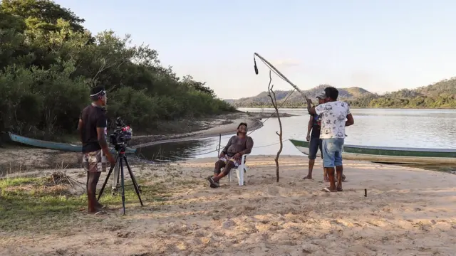 Fotografia colorida mostra um homem indígena sentadocomo sacar o bonus da arbetyuma cadeira na beira do rio sendo filmado por três outros homens indígenas