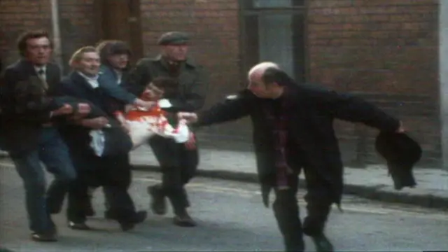 Un sacerdote agita un pañuelo manchado de sangre mientras cuatro hombres llevan a un hombre herido y ensangrentado por las calles de Londonderry durante The Troubles.