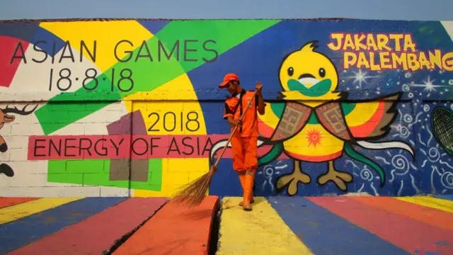 Petugas kebersihan menyapu dengan latar belakang mural Asian Games 2018 di Kampung Tematik, Kemayoran, Jakarta.