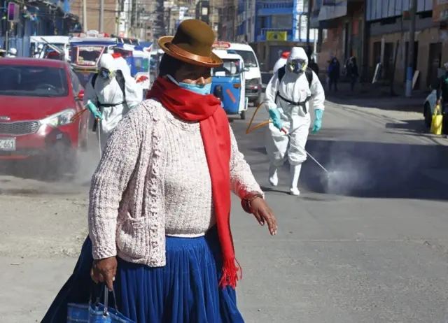 A woman in typical Aymara indigenous attire walks near municipal workers spraying disinfectant at a market in Puno, Peru