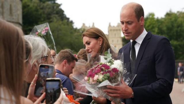 Foto colorida mostra o príncipe William, um homem branco adulto com cabelo rareando, ejogo b1betesposa Kate, uma mulher brancajogo b1betcabelo castanho comprido, recebendo floresjogo b1betdiversas pessoas reunidas atrásjogo b1betuma barreira