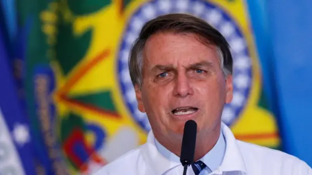 Bolsonaro fala ao microfone, com bandeira atrás