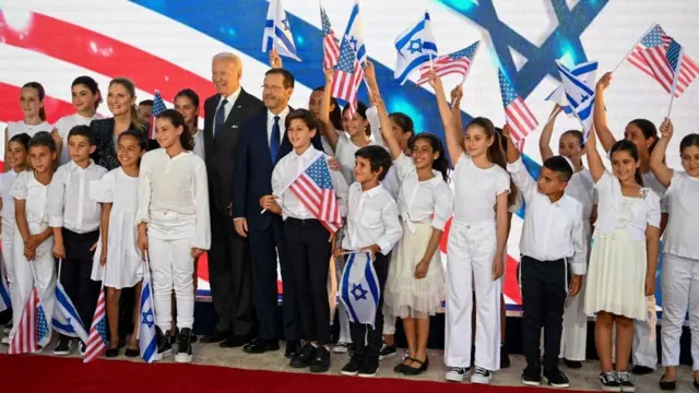 جو بایدن در کنار رئیس‌جمهور اسرائیل و کودکان اسرائیلی که پرچم‌های دو کشور را در دست دارند