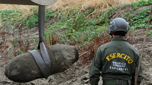 Um homem com capacete e uniforme verde no qual está escrito exércitobrasileirao 2024 tabelaitaliano observa uma bomba com aspecto antigo ser levantada por uma máquina não visível na margembrasileirao 2024 tabelaum rio