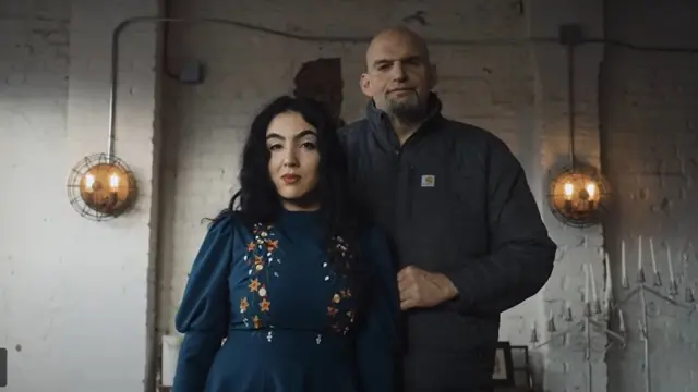 Fotografia colorida mostra mulher brasileirapele branca e cabelo preto, usando um vestido verde escuro, ao ladoum homem branco, alto, careca ebarba