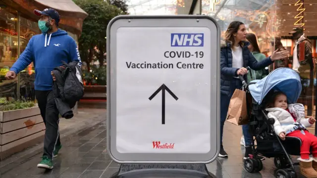 People passing by the NHS covid-19 vaccination Centre direction sign in Westfield Shopping Centre.