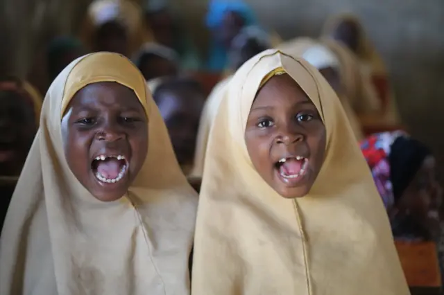 Deux filles auxquelles il manque des dents de lait ouvrent grand la bouche en récitant dans une salle de classe.