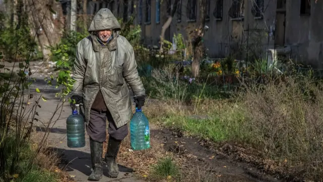 Hombre que lleva dos botellas grandes de agua camina por un sendero frente a un edificio dañado.