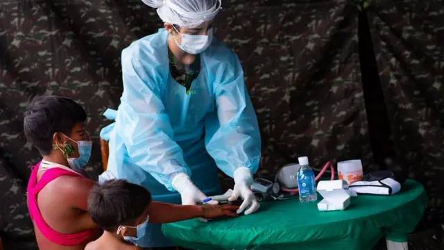 Com pano camuflado ao fundo, enfermeira prepara doses, ao lado de adulto e criança indígenas sentados - todos de máscara