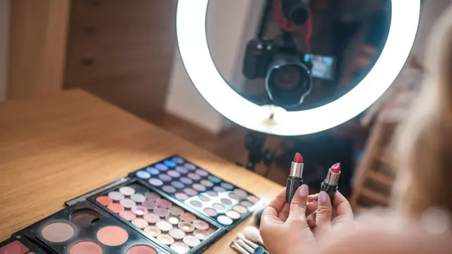 Young vlogger, vlogging about make-up - we see, over her shoulder, the camera, the ring-flash, and make-up on the table. She is holding two different colour lipsticks