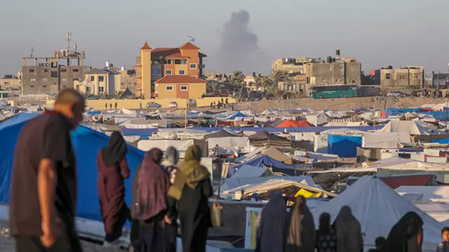 Smoke rises in the distance while people stand in front of a tent community in Rafah on 7 May