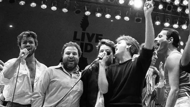 Michael (à esquerda) esteve no palco na despedida final do Live Aid, ao lado do promotor do evento Harvey Goldsmith e dos músicos Bono, Paul McCartney, Bob Geldof e Freddie Mercury.