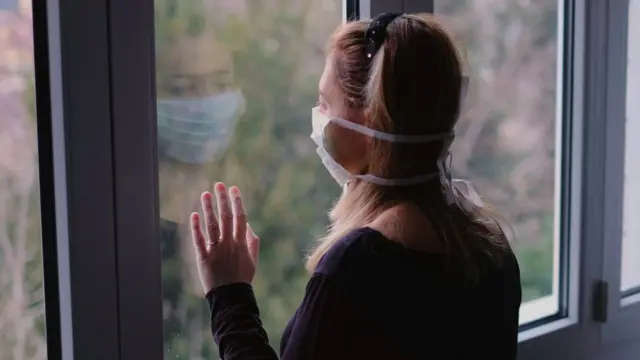 Una sola mujer con una mascarilla mira afuera por una ventana