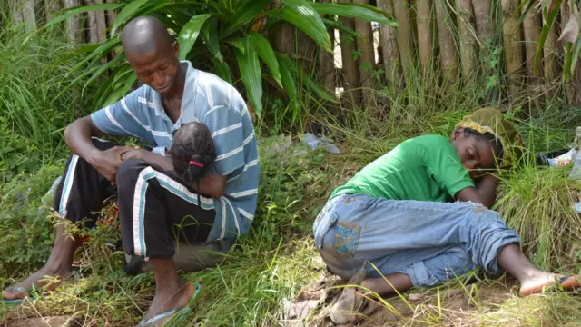 Un padre con su niña pequeña en brazos y su mujer enferma recostada en el pasto aguardan para ser atendidos en una clínica
