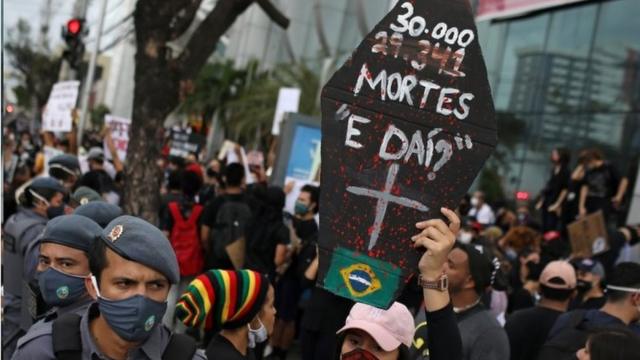 Em protesto, com policiais e manifestantes de máscara, pessoa levanta cartaz em formato de caixão e frase: 30 mil mortes, e daí?