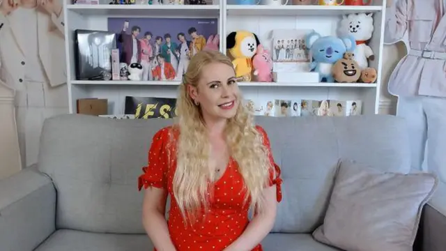 Jenny has long wavy blonde hair. She's wearing a red spotty dress and is sat on a grey sofa. In the background there is a book shelf full of soft cuddly toys and a cardboard cutout of K-Pop boyband.